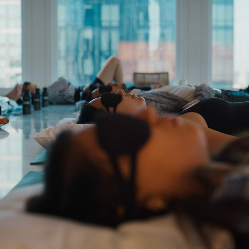 Group of people lying on yoga mats with eye masks in a bright room, relaxing near large city-view windows.