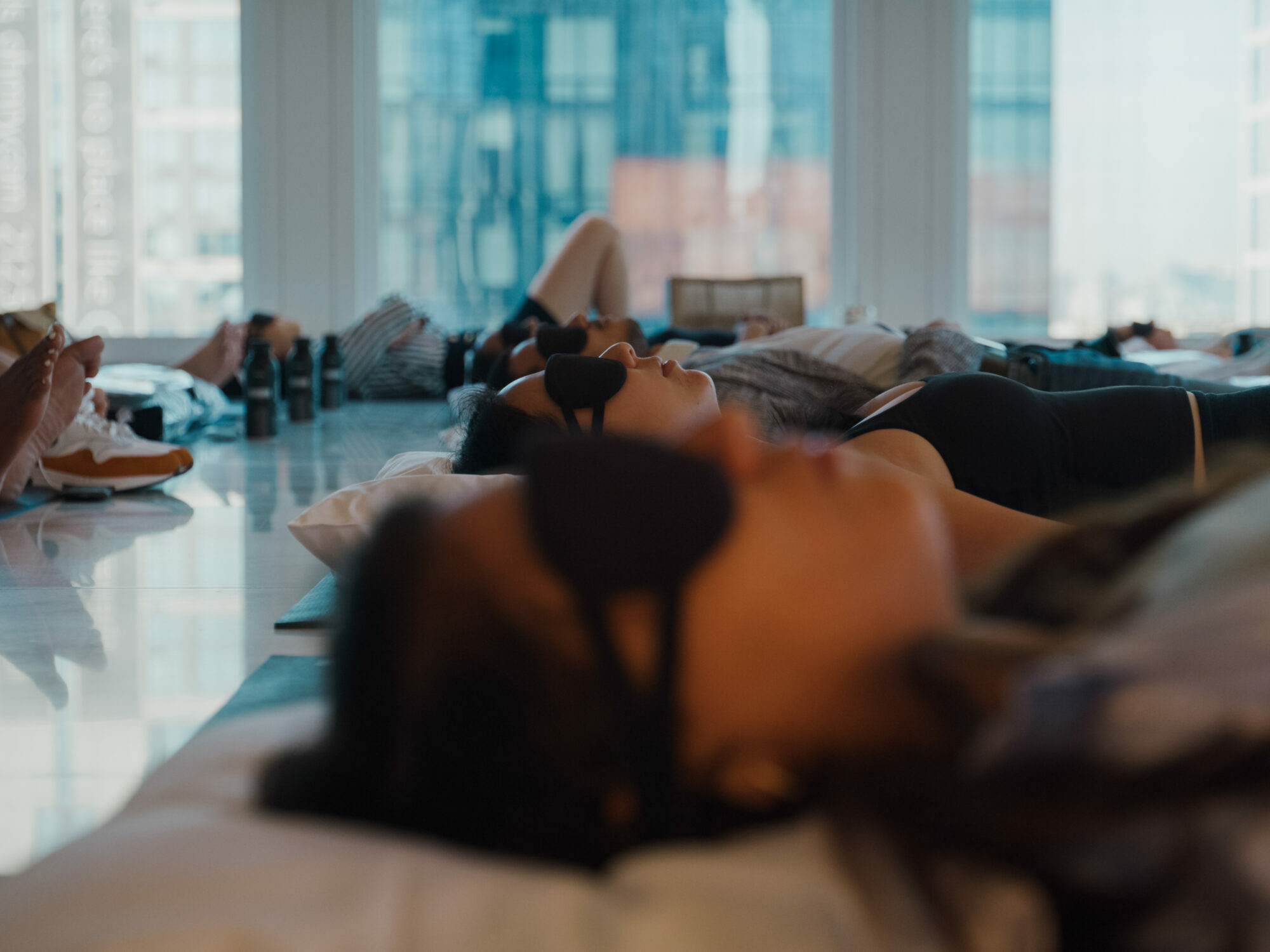 Group of people lying on yoga mats with eye masks in a bright room, relaxing near large city-view windows.