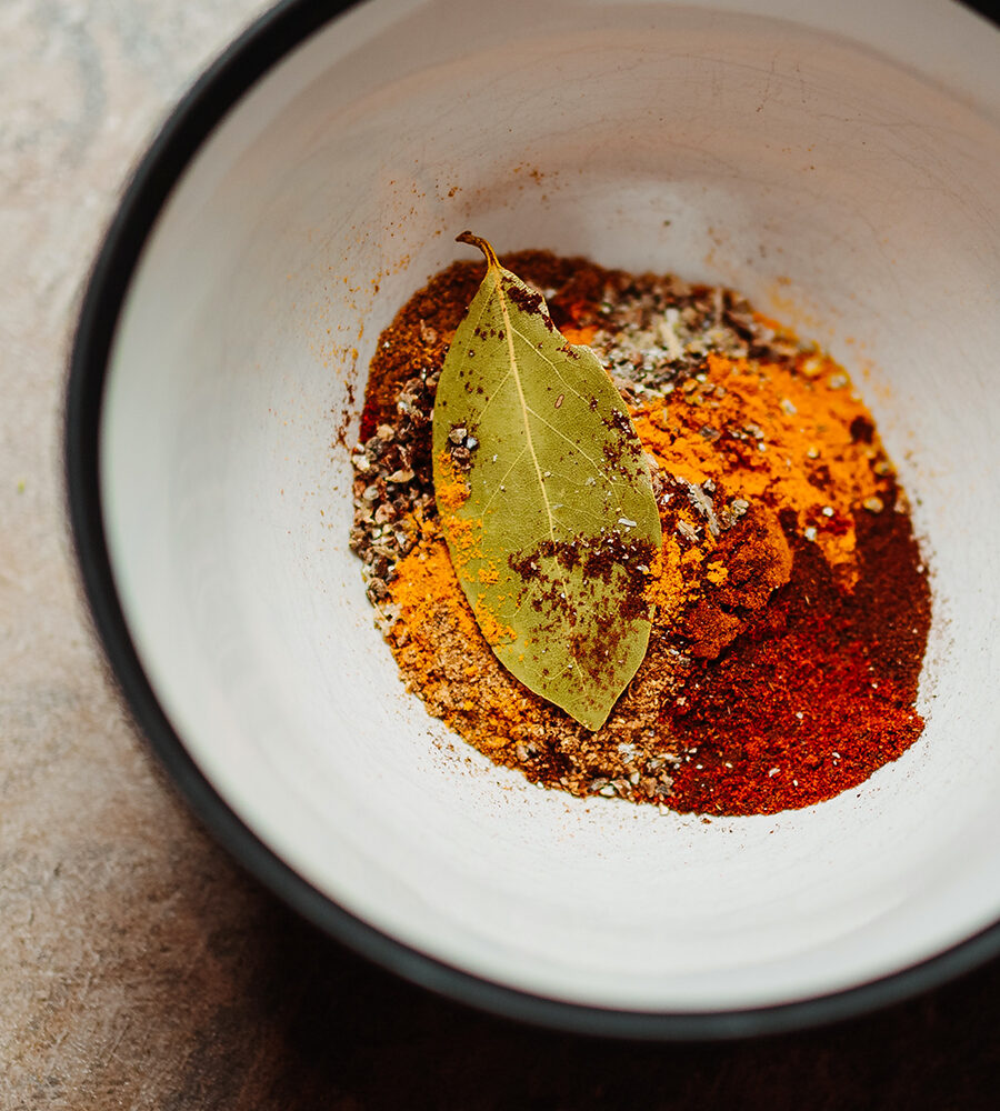 White bowl with black rim holding colorful ground spices—turmeric, paprika, cumin, pepper—and a dried bay leaf on top.