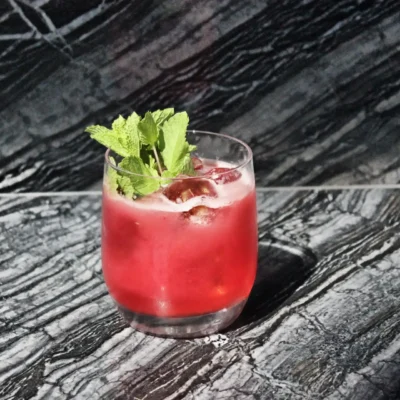 Red cocktail with ice and mint in short glass on dark marbled surface.