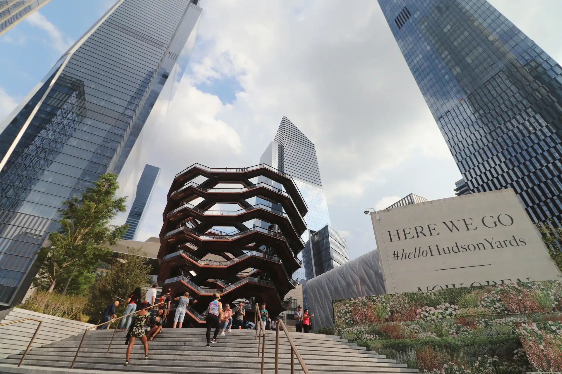 View of the Vessel surrounded by skyscrapers, with people walking and sign reading “HERE WE GO #helloHudsonYards”