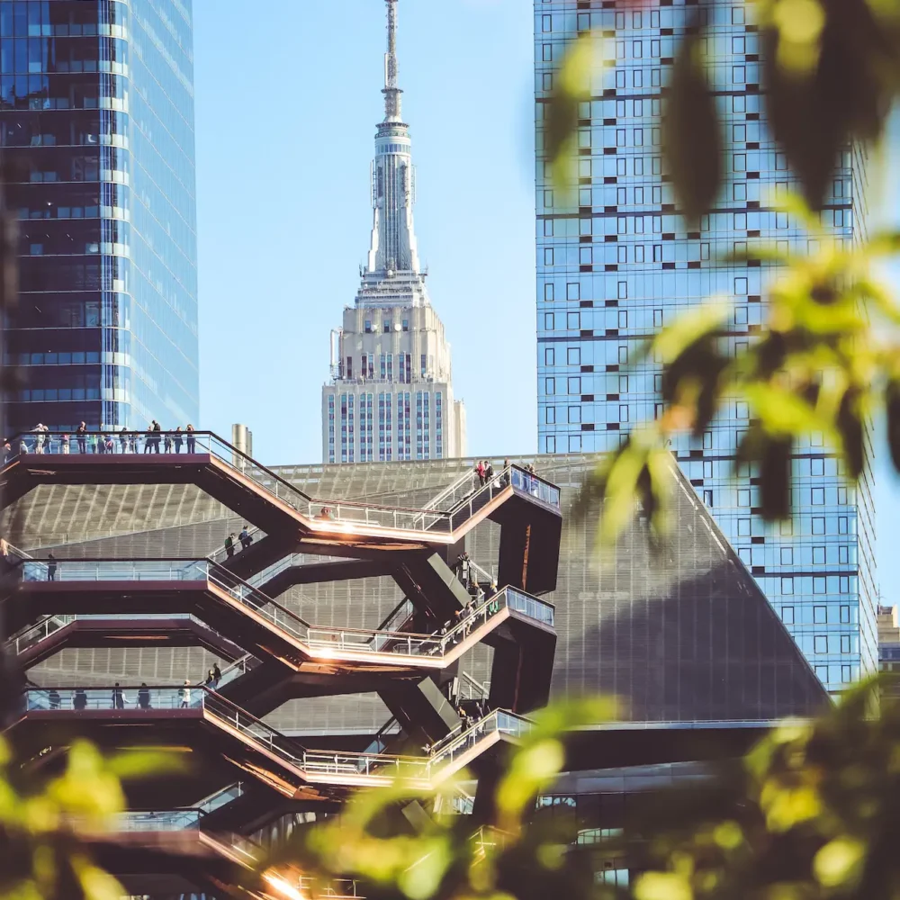 Empire State Building seen between modern towers and Vessel staircases, with foliage adding depth to NYC skyline