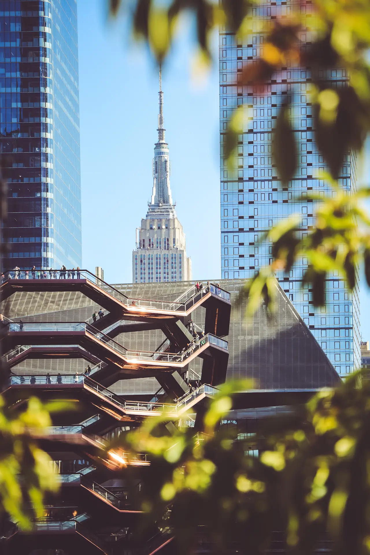 Empire State Building seen between modern towers and Vessel staircases, with foliage adding depth to NYC skyline