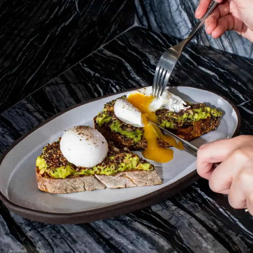 Close-up of avocado toast with poached eggs on a dark marble surface at Electric Lemon, New York City, with yolk spilling as a person cuts into the toast with knife and fork, capturing a refined brunch moment of flavor and craftsmanship.