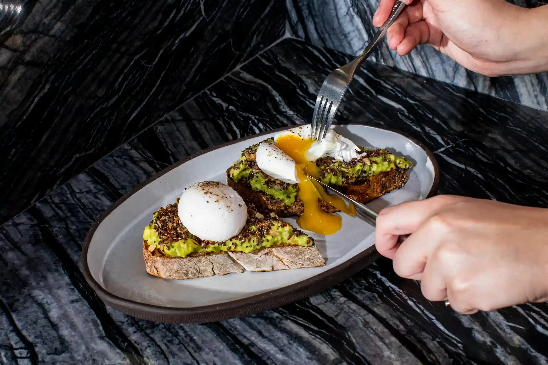 Close-up of avocado toast with poached eggs on a dark marble surface at Electric Lemon, New York City, with yolk spilling as a person cuts into the toast with knife and fork, capturing a refined brunch moment of flavor and craftsmanship.