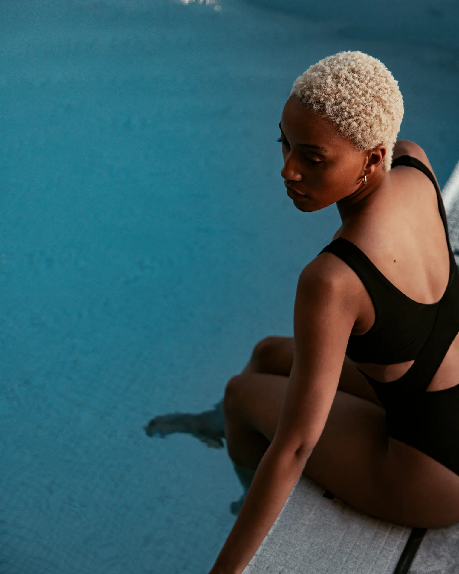 Person in black swimsuit sitting poolside with feet in water, looking over shoulder at clear blue pool.