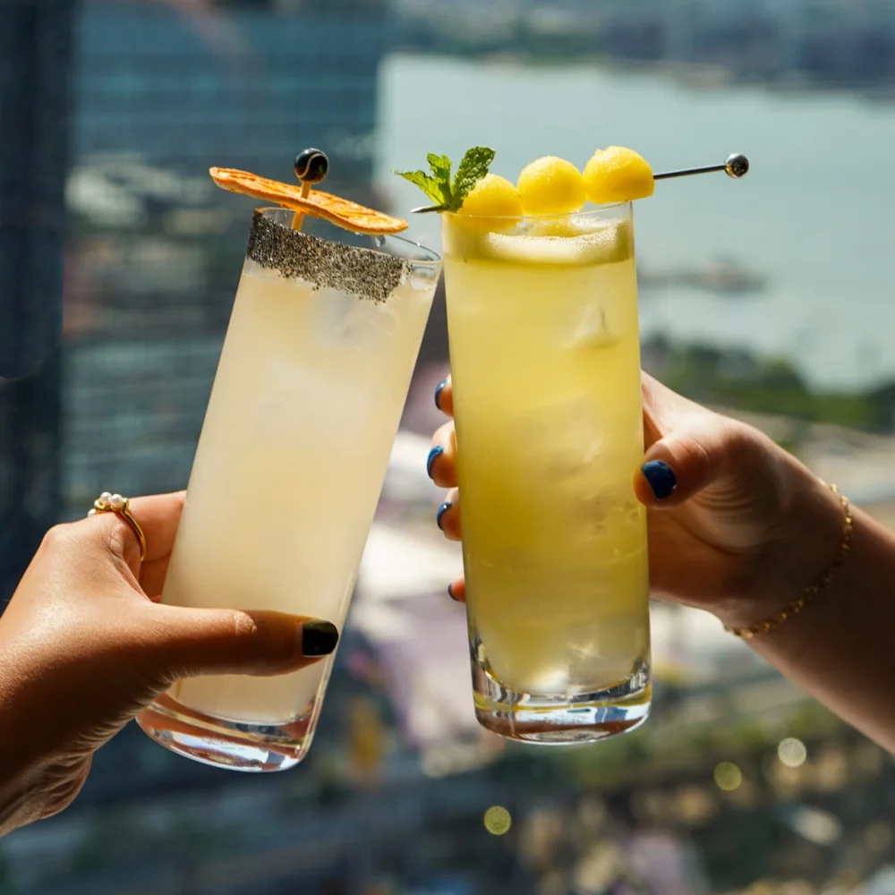 Two hands holding cocktails with citrus and melon garnishes against blurred city and waterfront backdrop