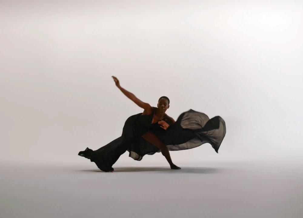Dancer in flowing black outfit mid-motion against light backdrop, expressing strength and grace