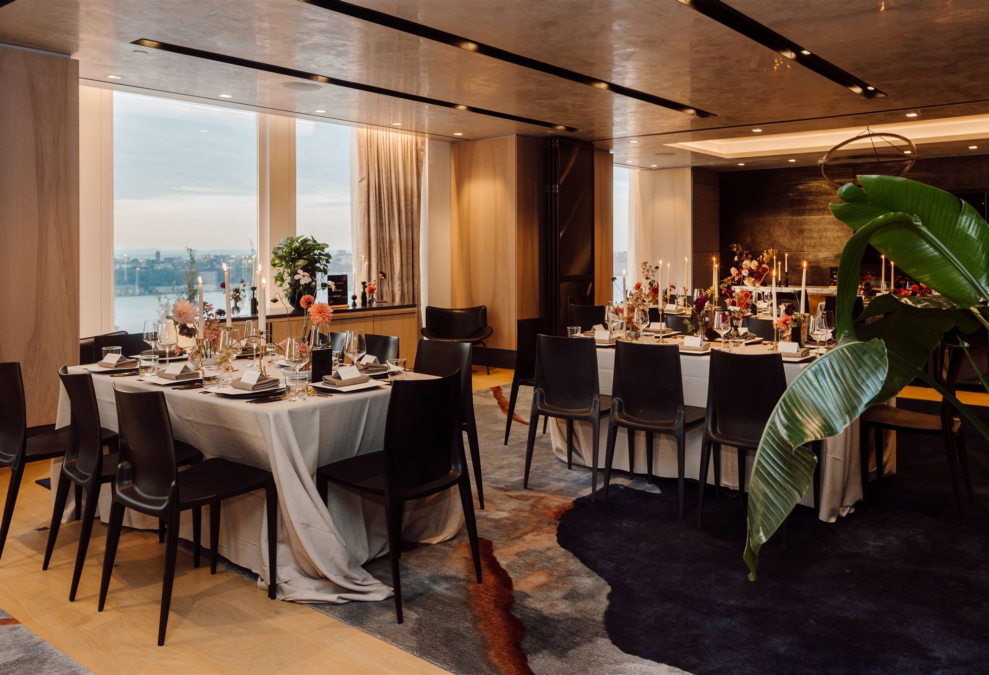 Formal dining setup with white tablecloths, floral centerpieces, and city and waterfront views through large windows