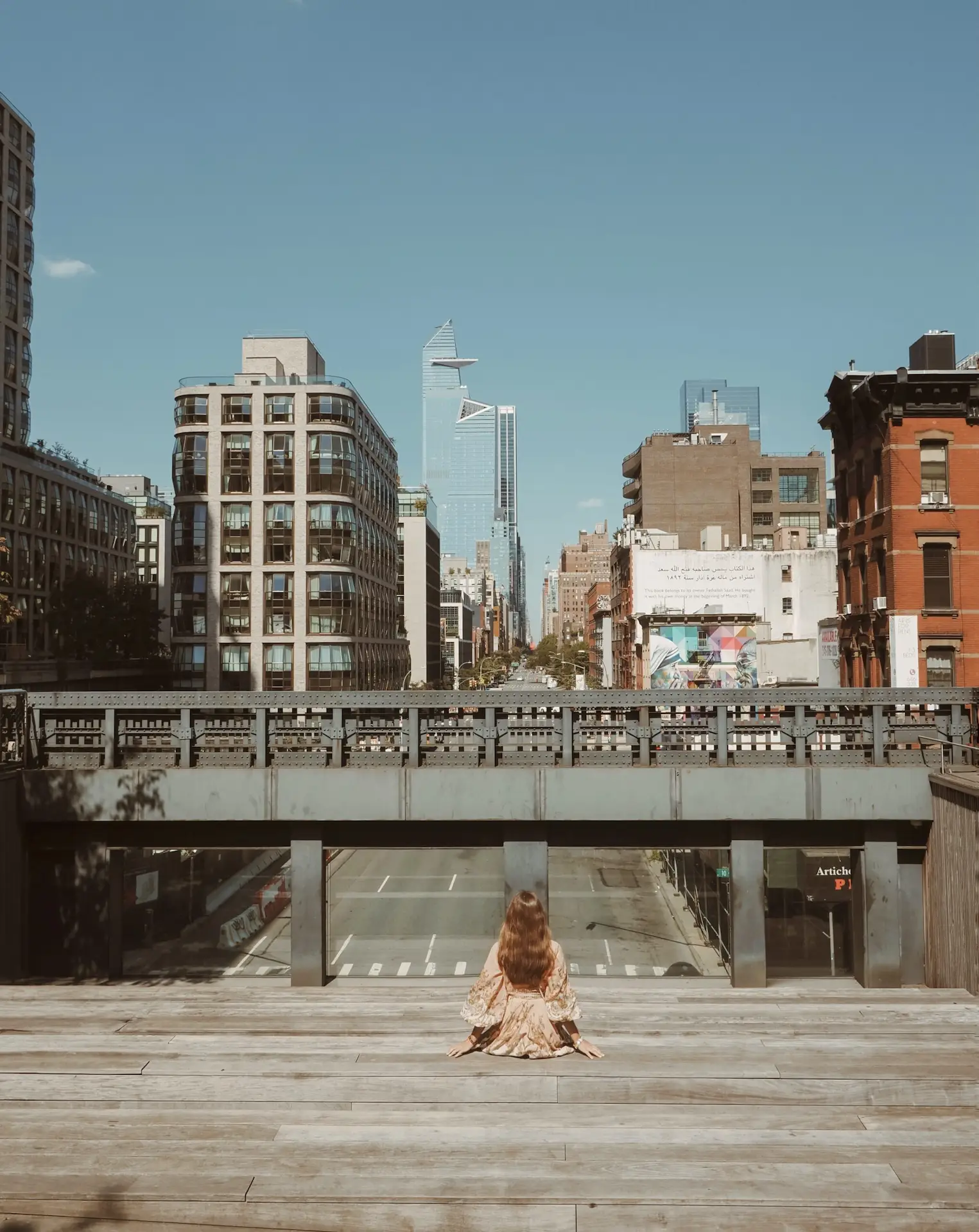 Person in patterned dress seated alone on wooden platform overlooking city skyline with mix of architecture