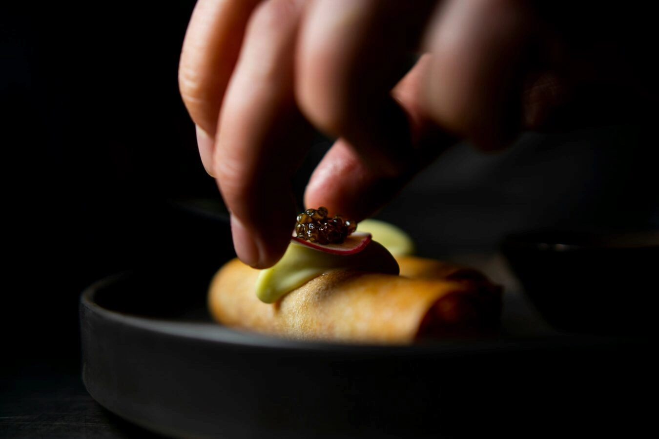 Close-up of hand placing caviar garnish on gourmet roll with radish and green vegetable on dark plate