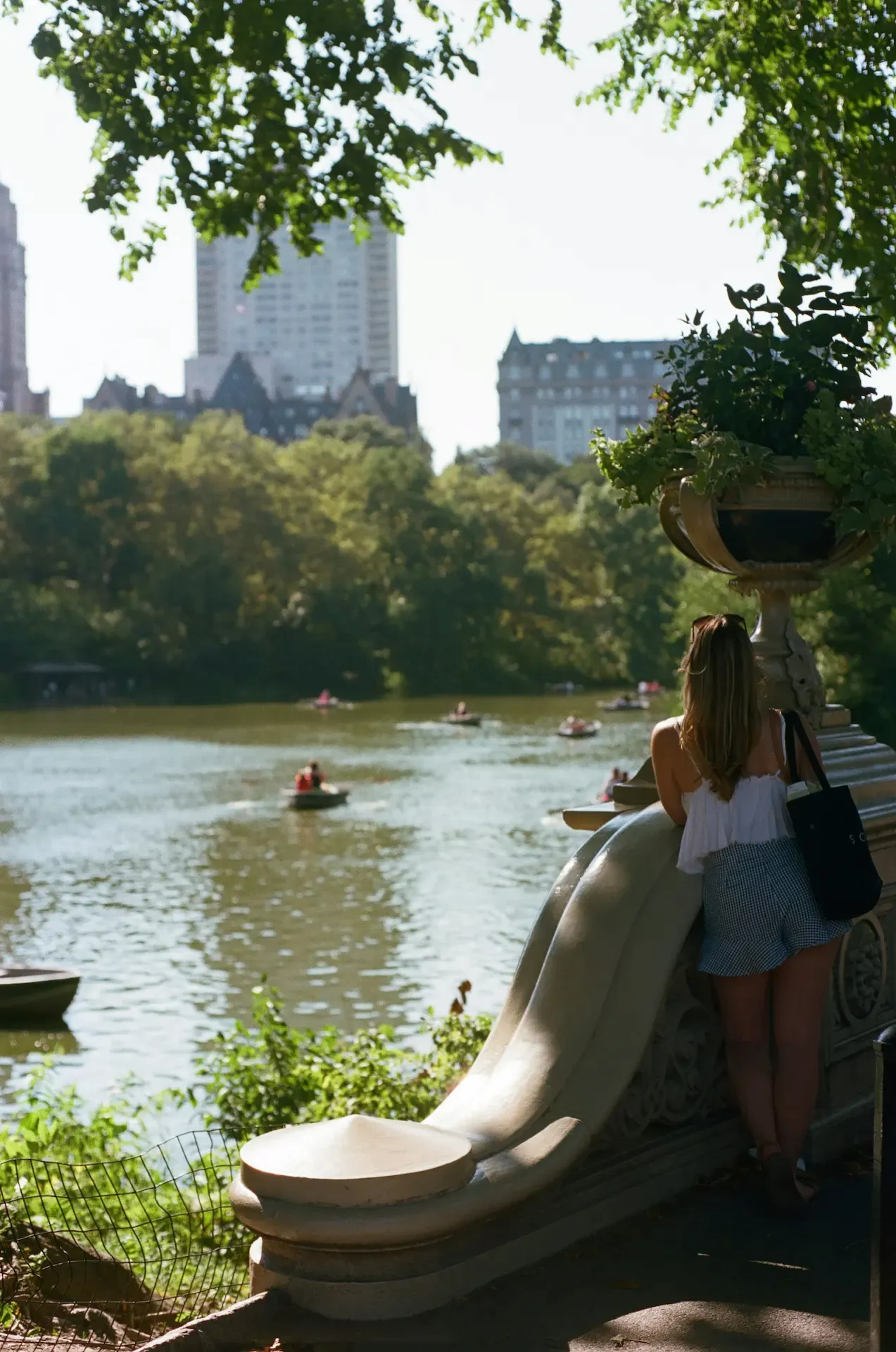 Person standing on a stone railing overlooking a lake with rowboats in Central Park, surrounded by trees & city buildings.