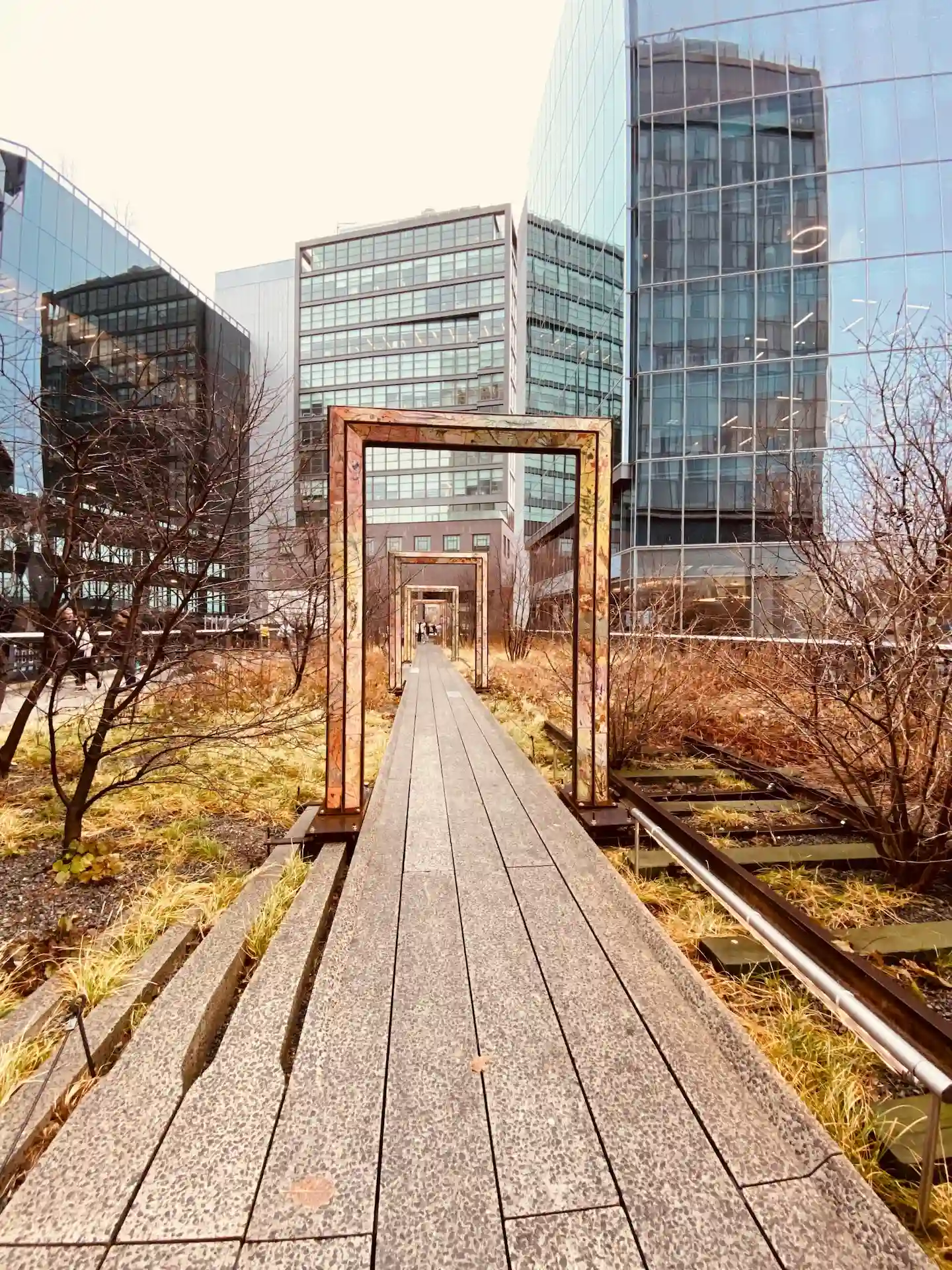 Elevated walkway with rust-colored arches and dry plants surrounded by glass buildings in urban park