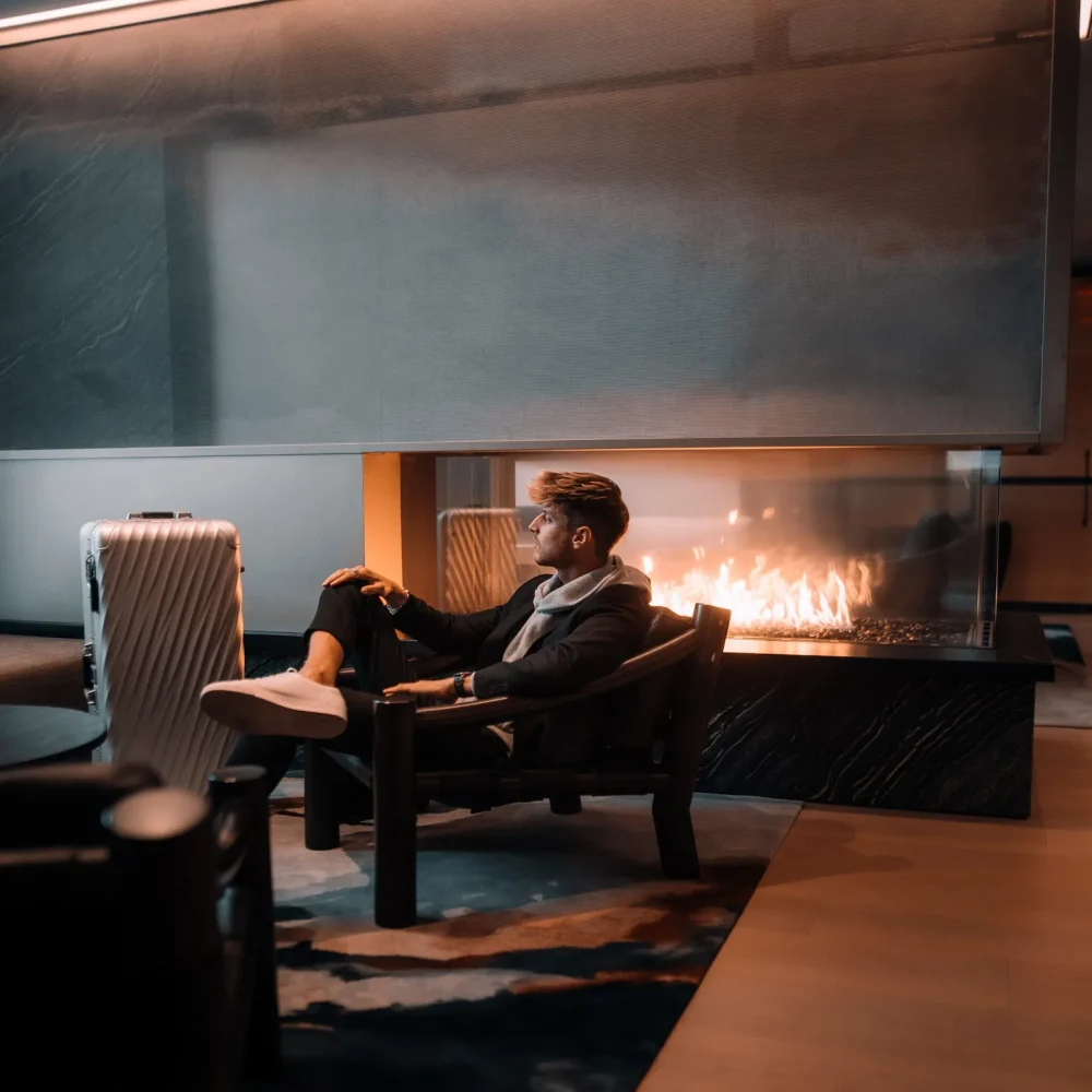 Person in dark suit seated by sleek fireplace at Equinox hotel lounge with suitcase and warm lighting