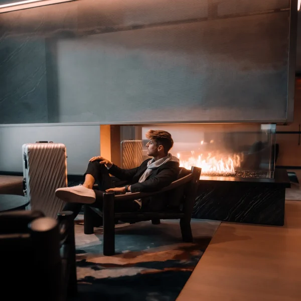 Person in dark suit seated by sleek fireplace at Equinox hotel lounge with suitcase and warm lighting