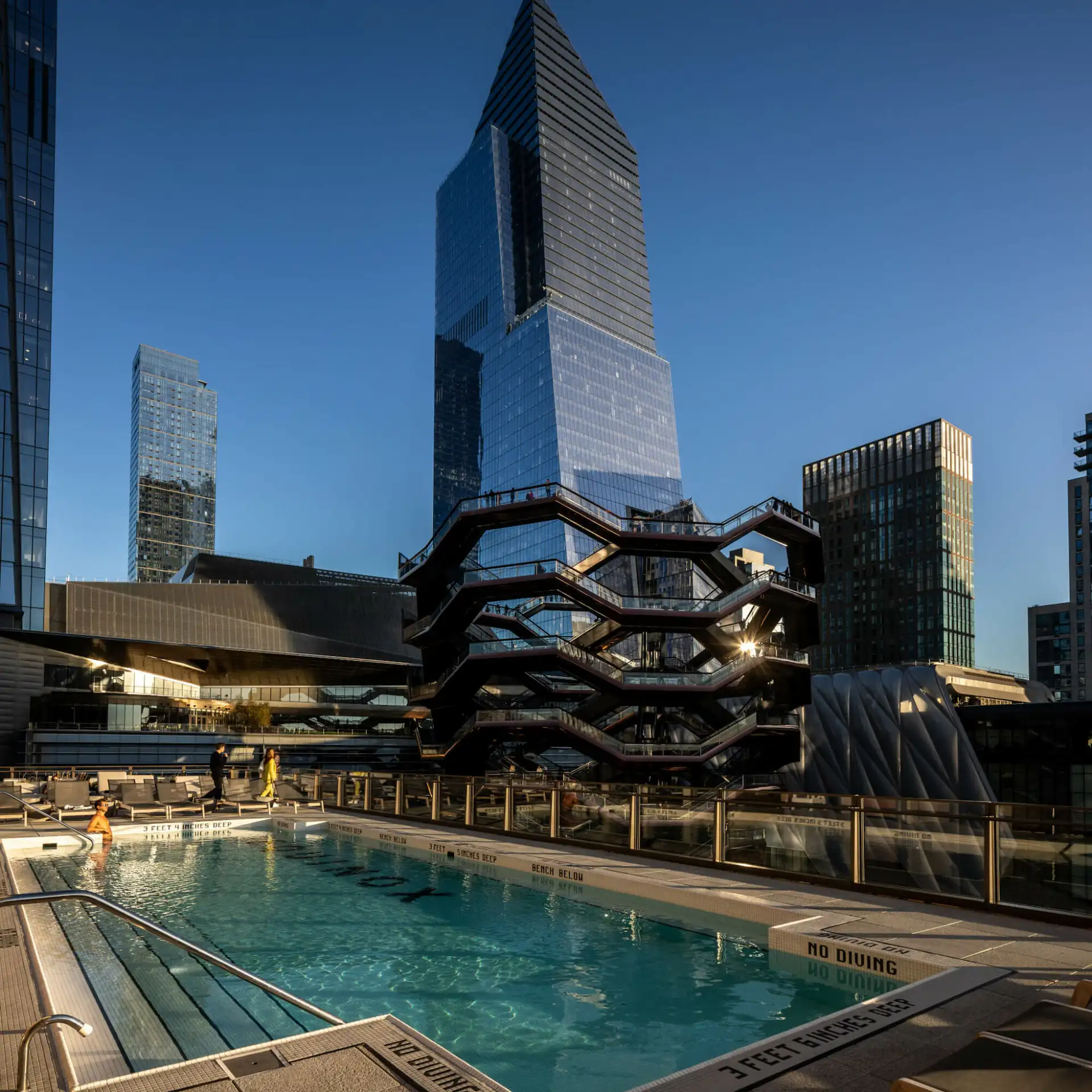 Rooftop pool overlooking modern skyscrapers and the Vessel structure in New York City at sunset.