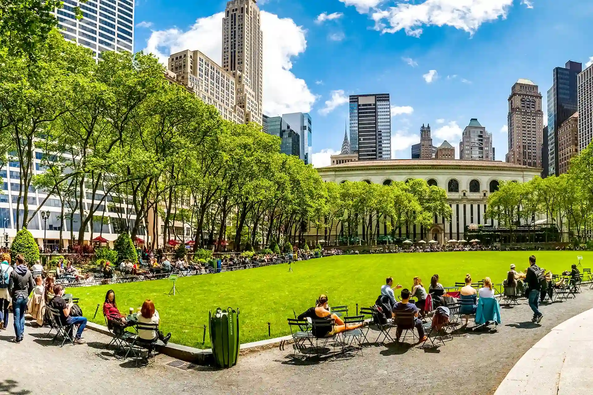 Bryant Park on sunny day with people relaxing on lawn and chairs, surrounded by trees and skyscrapers