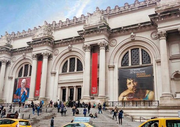 The Met facade with neoclassical columns, red banners, exhibition posters, and visitors on museum steps