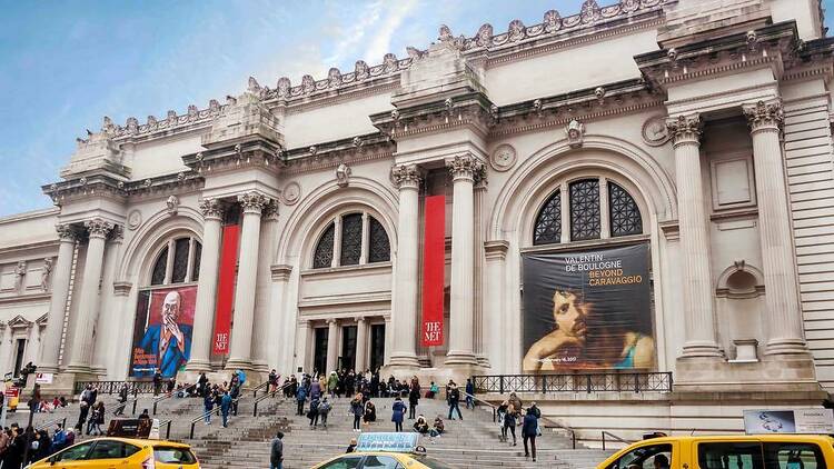The Met facade with neoclassical columns, red banners, exhibition posters, and visitors on museum steps