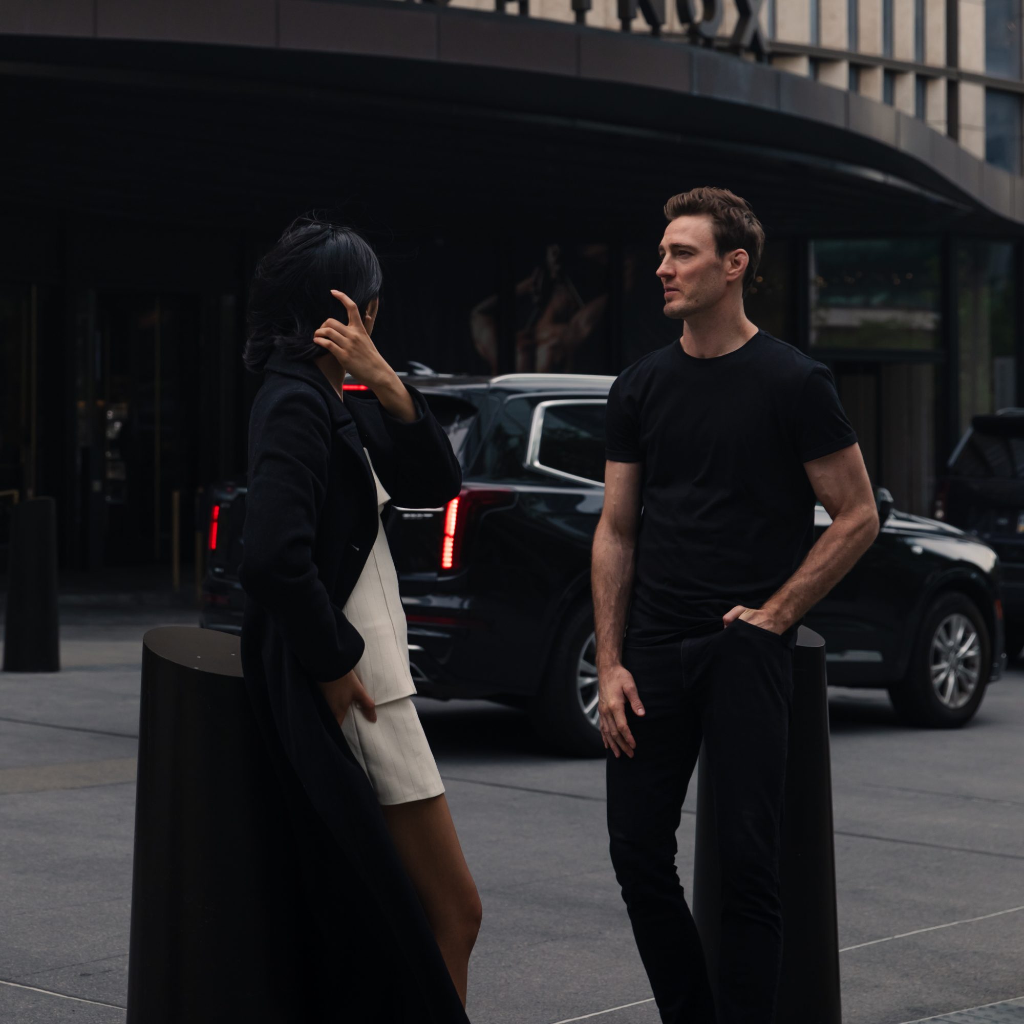 Two people talking on a city sidewalk beside a black luxury SUV in front of a modern glass building.