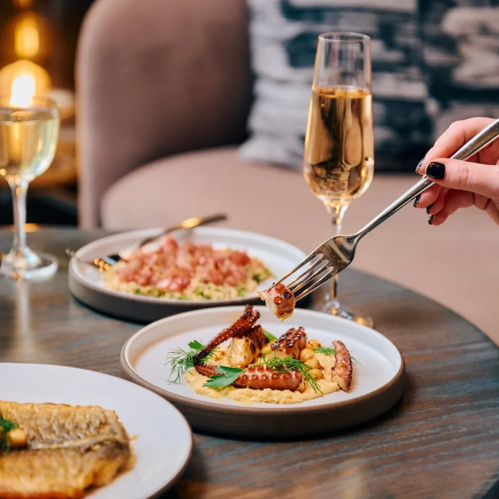 A diner uses a fork to eat grilled octopus at Electric Lemon, with glasses of wine and various small plates on the table.