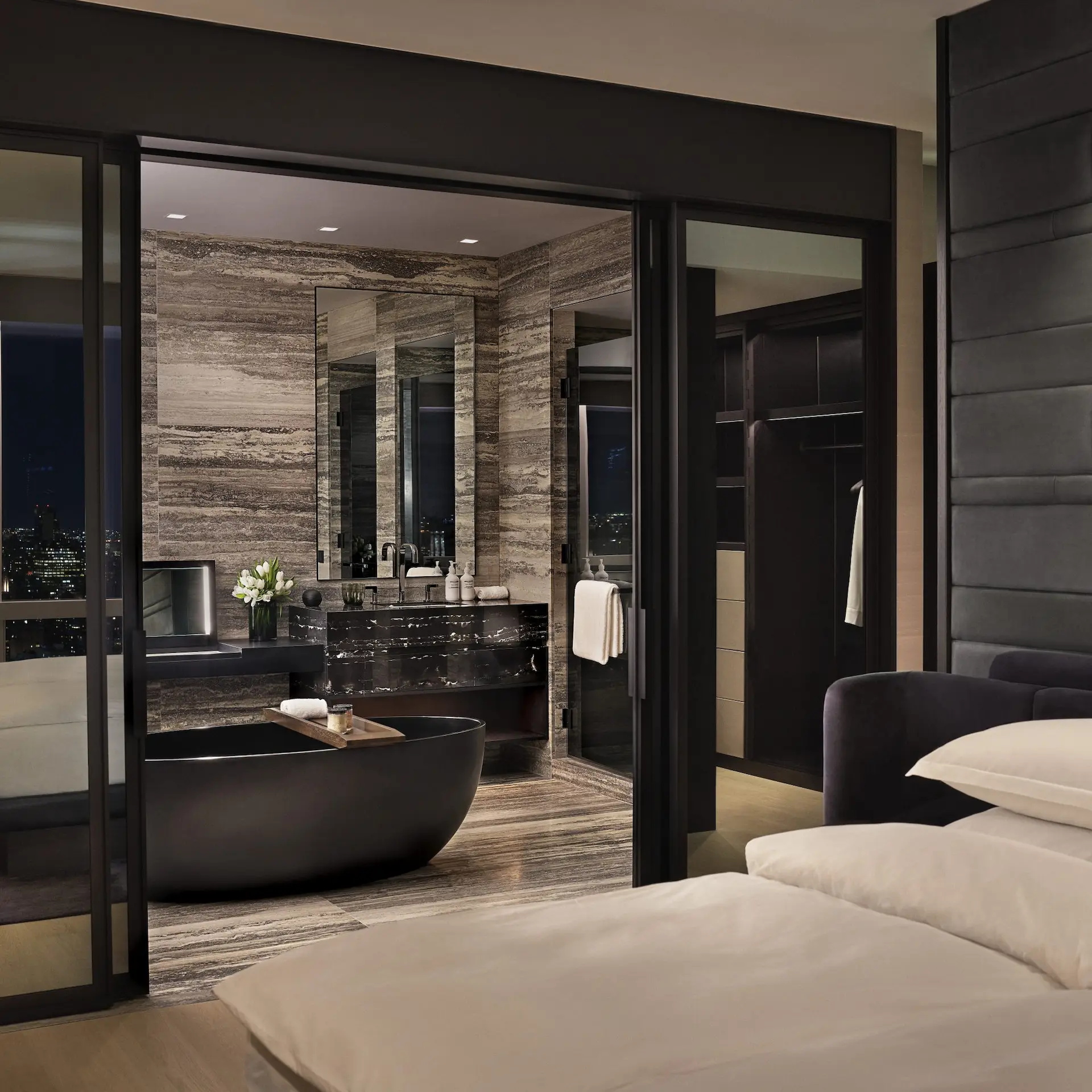 Modern suite with a bed facing a glass partition that reveals a bathroom with a black soaking tub and nighttime city light.