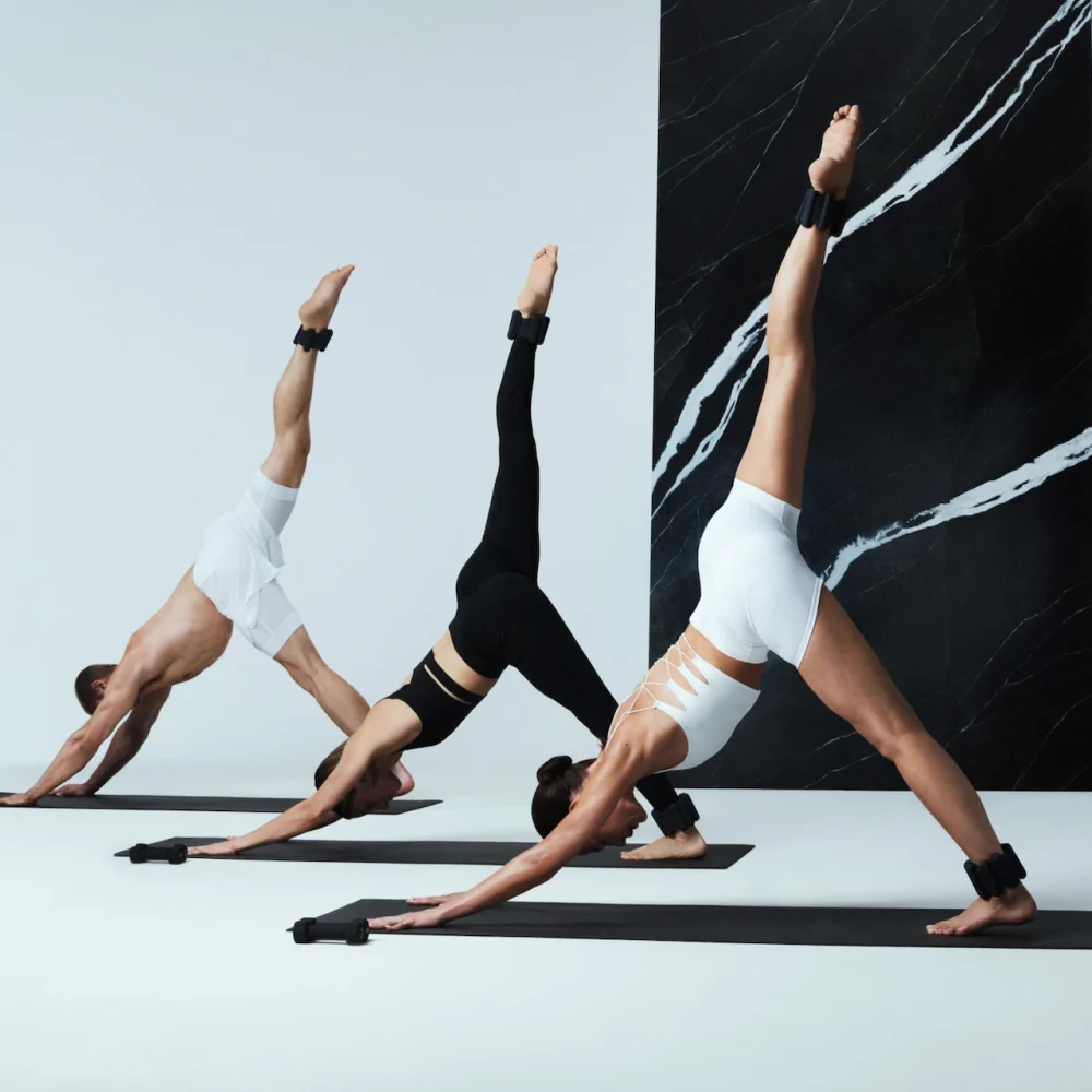 Three individuals in three-legged downward dog pose with ankle weights, set against minimalist studio backdrop, evoking strength, balance, and elevated movement
