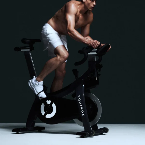 Individual riding Equinox-branded stationary bike in dark setting, evoking intensity, focus, and elevated cardio training