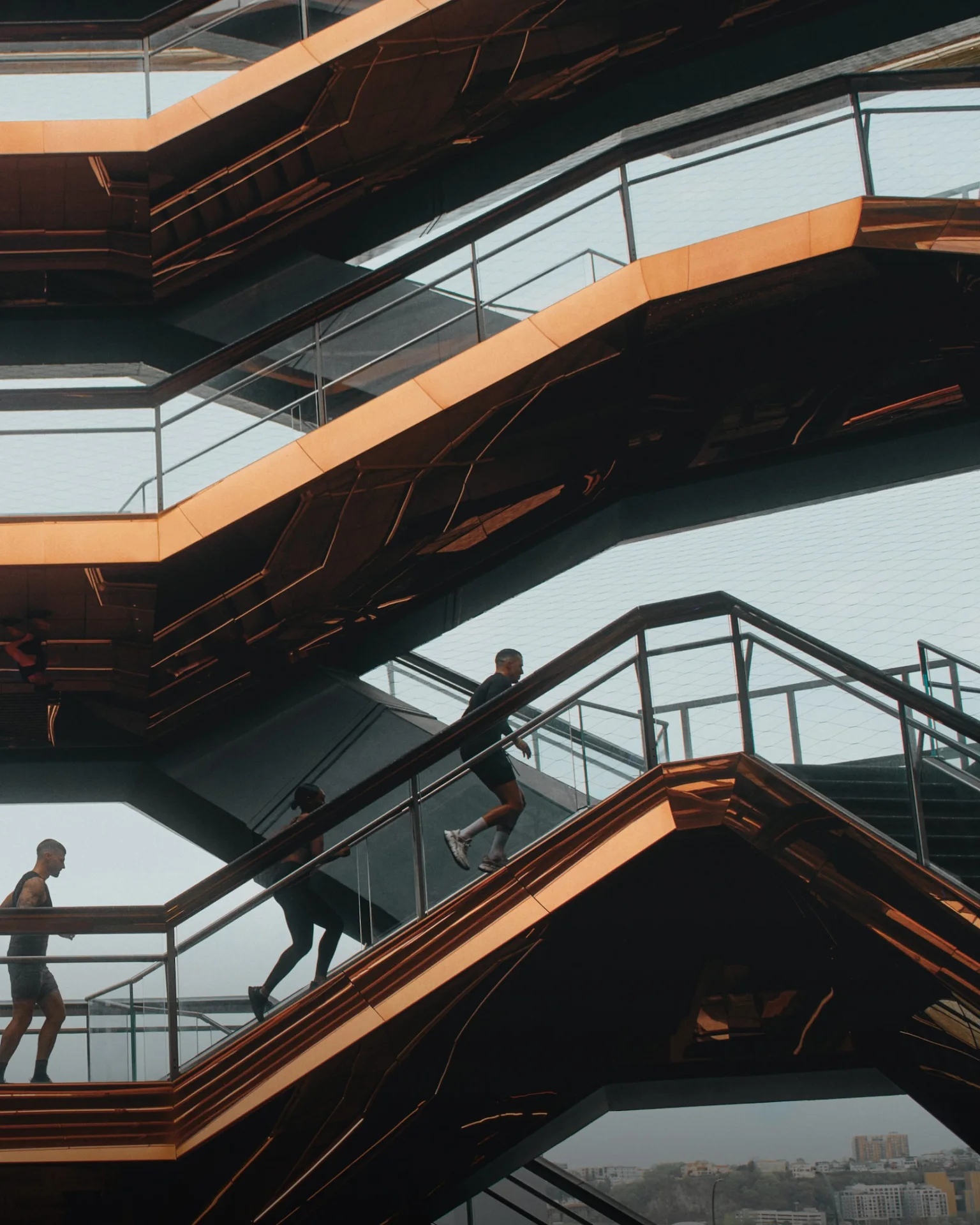 People walking up angular bronze‑edged staircases with glass railings in a architectural structure overlooking a cityscape.