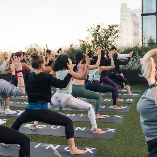 Group fitness class practicing yoga poses on rooftop mats with a city skyline in the background.