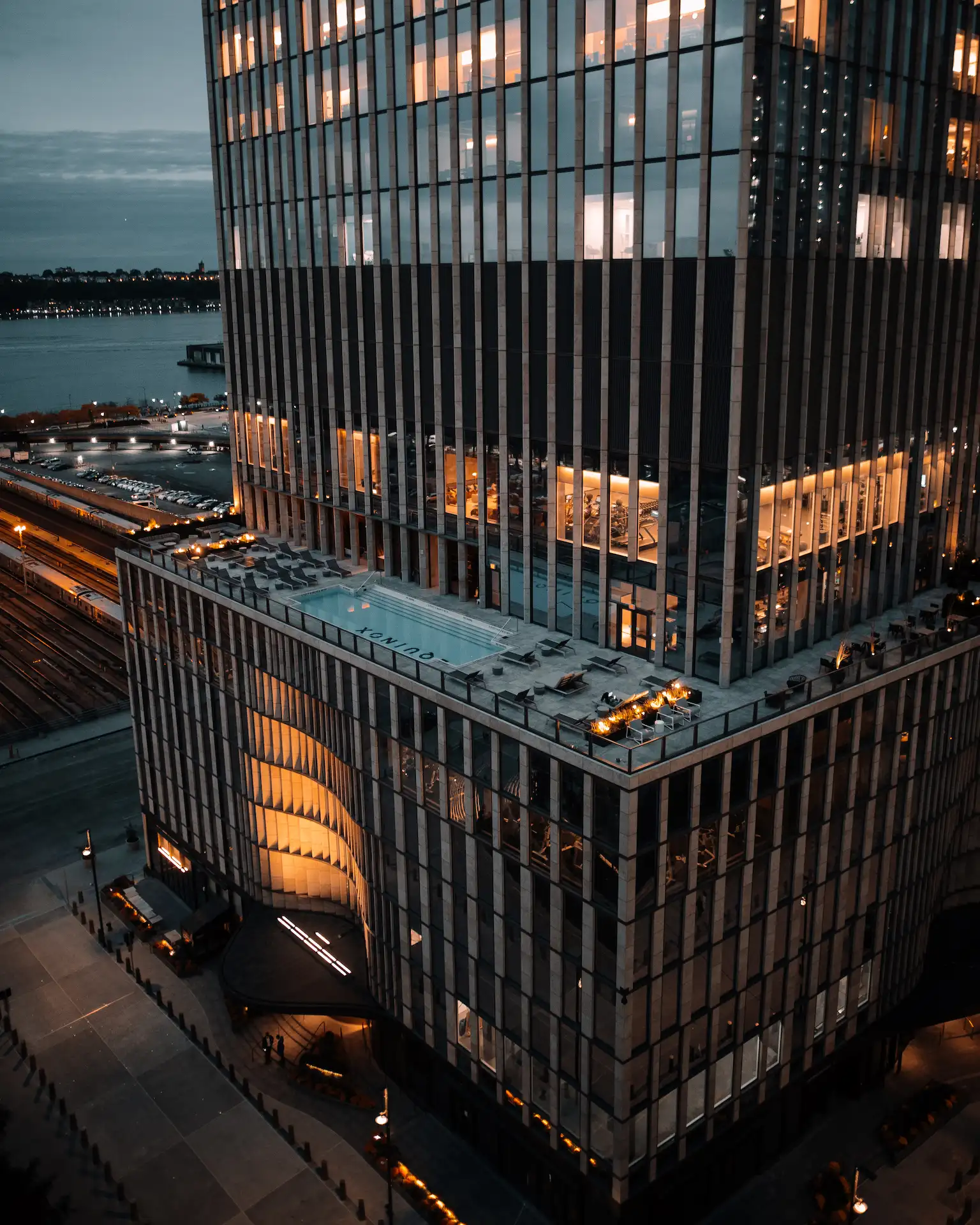 Modern high-rise building with illuminated terrace and glass facade, set against evening sky and urban waterfront, evoking upscale city living and architectural elegance