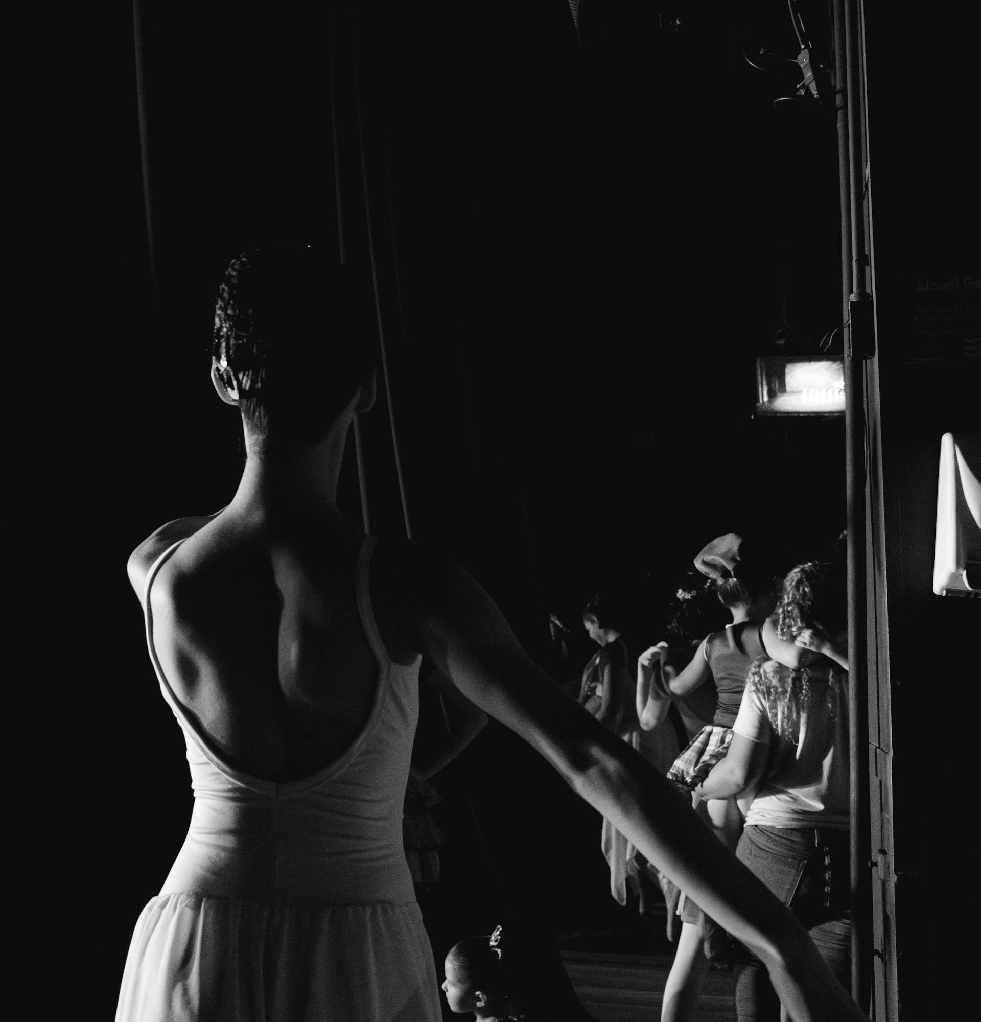 Ballet dancer backstage in low light, adjusting position while other performers prepare behind the curtain