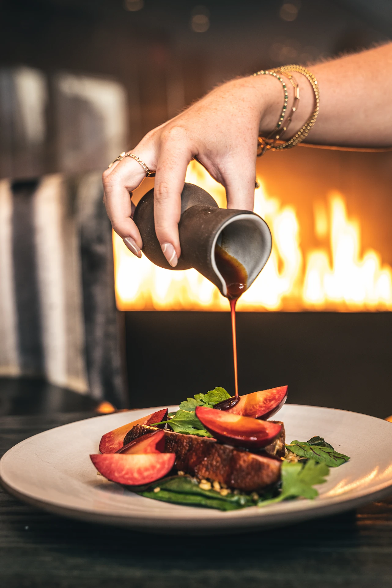 Hand pouring sauce over gourmet dish by a fireplace, evoking warmth, detail, and elevated dining ritual