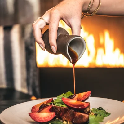 Hand pouring sauce over gourmet dish by a fireplace, evoking warmth, detail, and elevated dining ritual