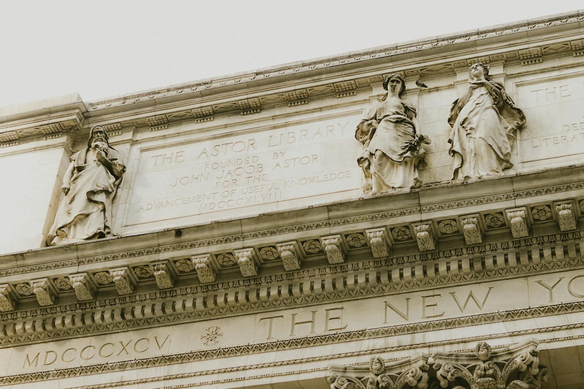 Detailed view of the New York Public Library facade showing sculpted classical figures, engraved ‘Astor Library’ inscription, and ornate stone cornice work against a pale sky.