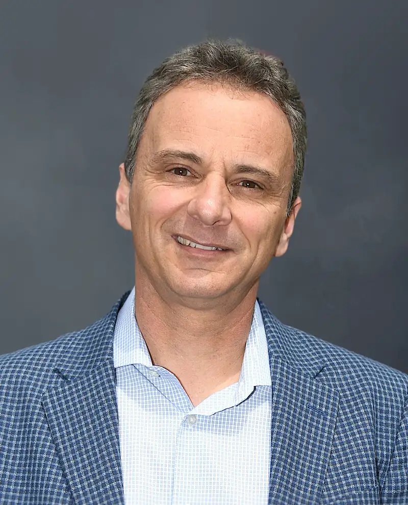 Professional portrait of a person wearing a light blue checkered blazer and collared shirt against a dark gray background.