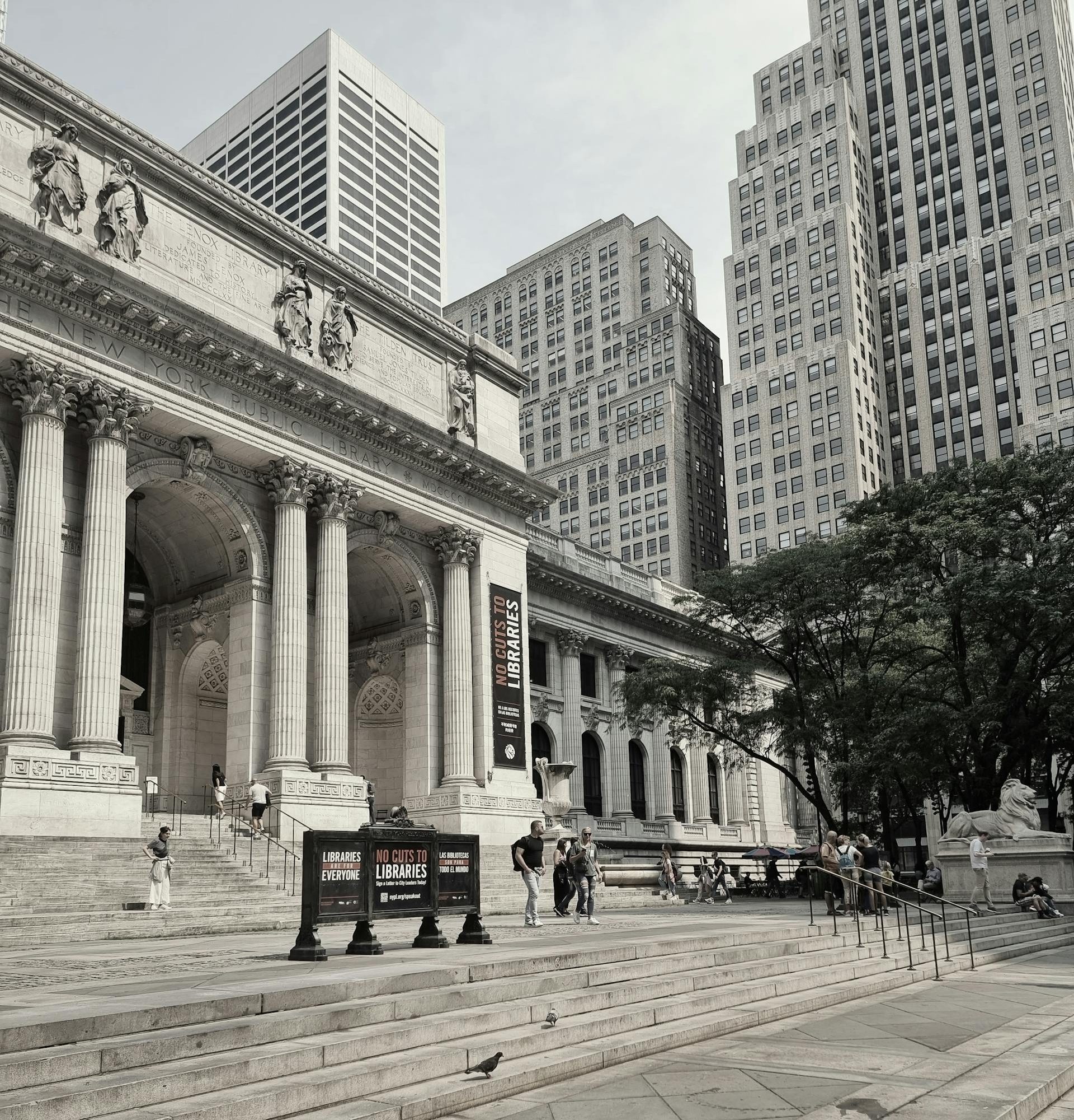 Exterior of the New York Public Library’s Stephen A. Schwarzman Building in Midtown Manhattan, featuring grand marble columns, wide steps, and people walking and sitting outside, with tall skyscrapers rising in the background.