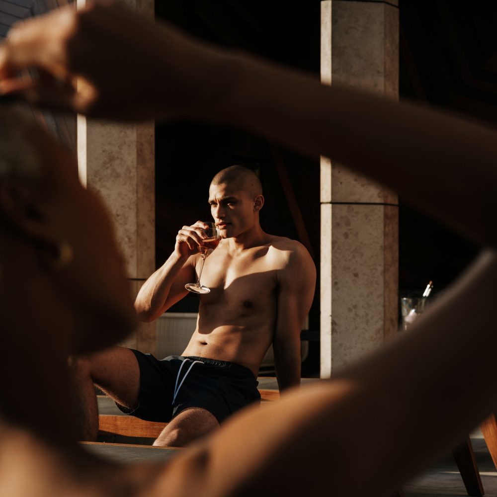 Two people relax poolside in warm sunlight while one sips a drink and another adjusts sunglasses in the foreground.