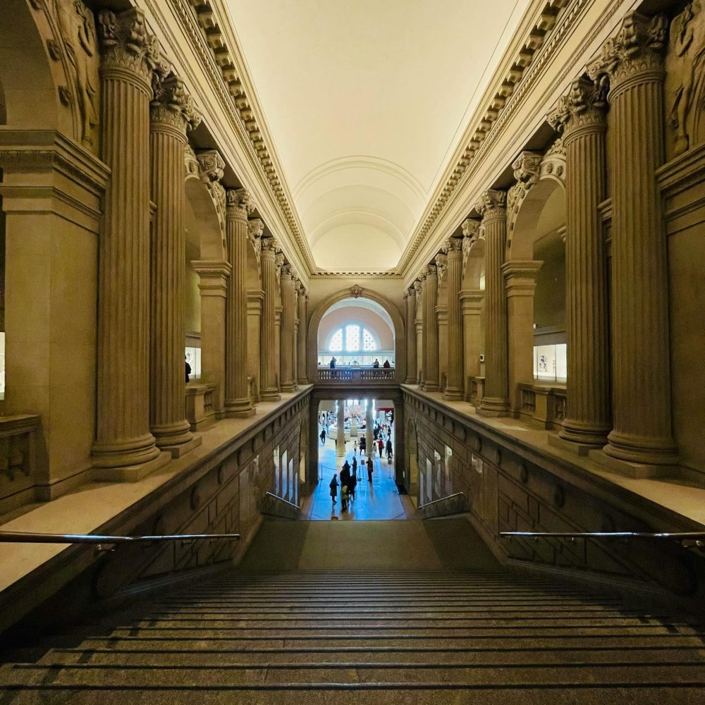 Grand staircase inside The Frick Collection in New York, featuring classical columns, vaulted ceilings, and visitors descending toward the main gallery level
