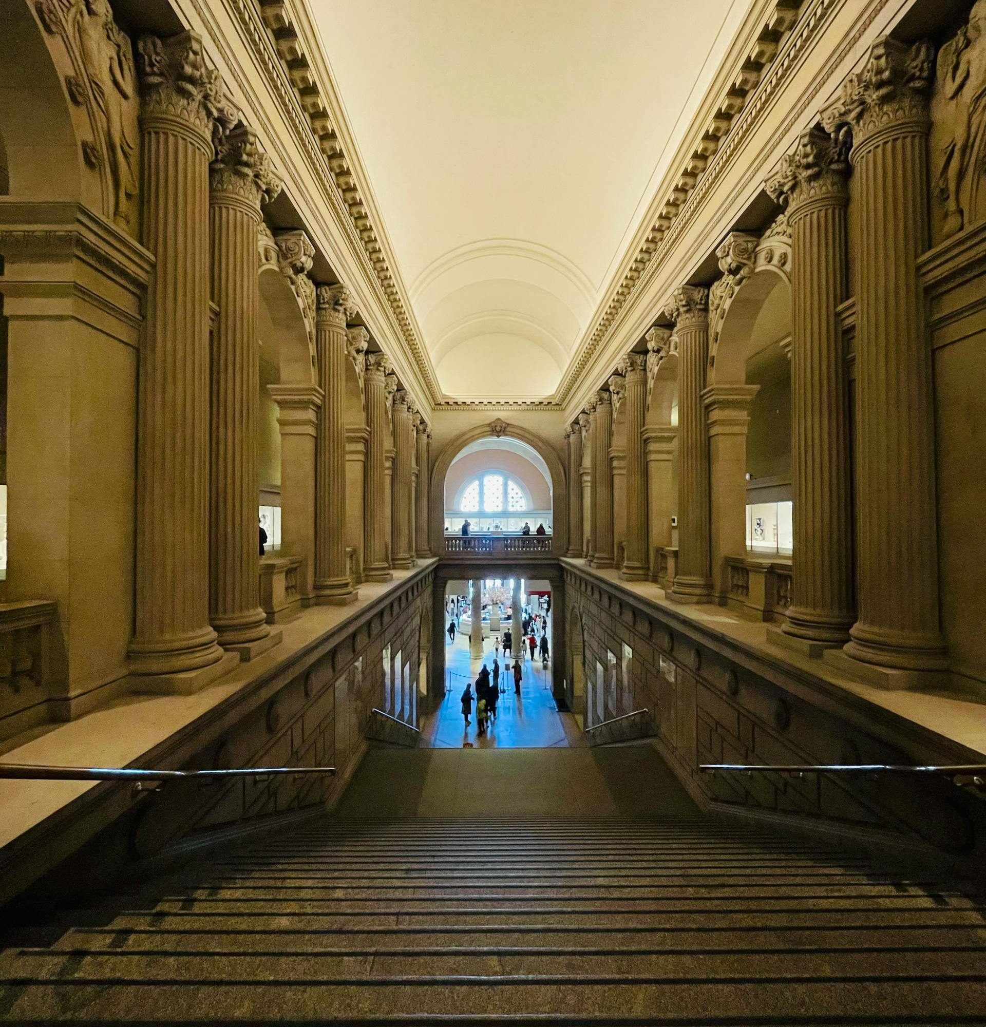 Grand staircase inside The Frick Collection in New York, featuring classical columns, vaulted ceilings, and visitors descending toward the main gallery level