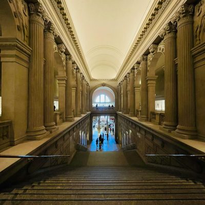 Grand staircase inside The Frick Collection in New York, featuring classical columns, vaulted ceilings, and visitors descending toward the main gallery level
