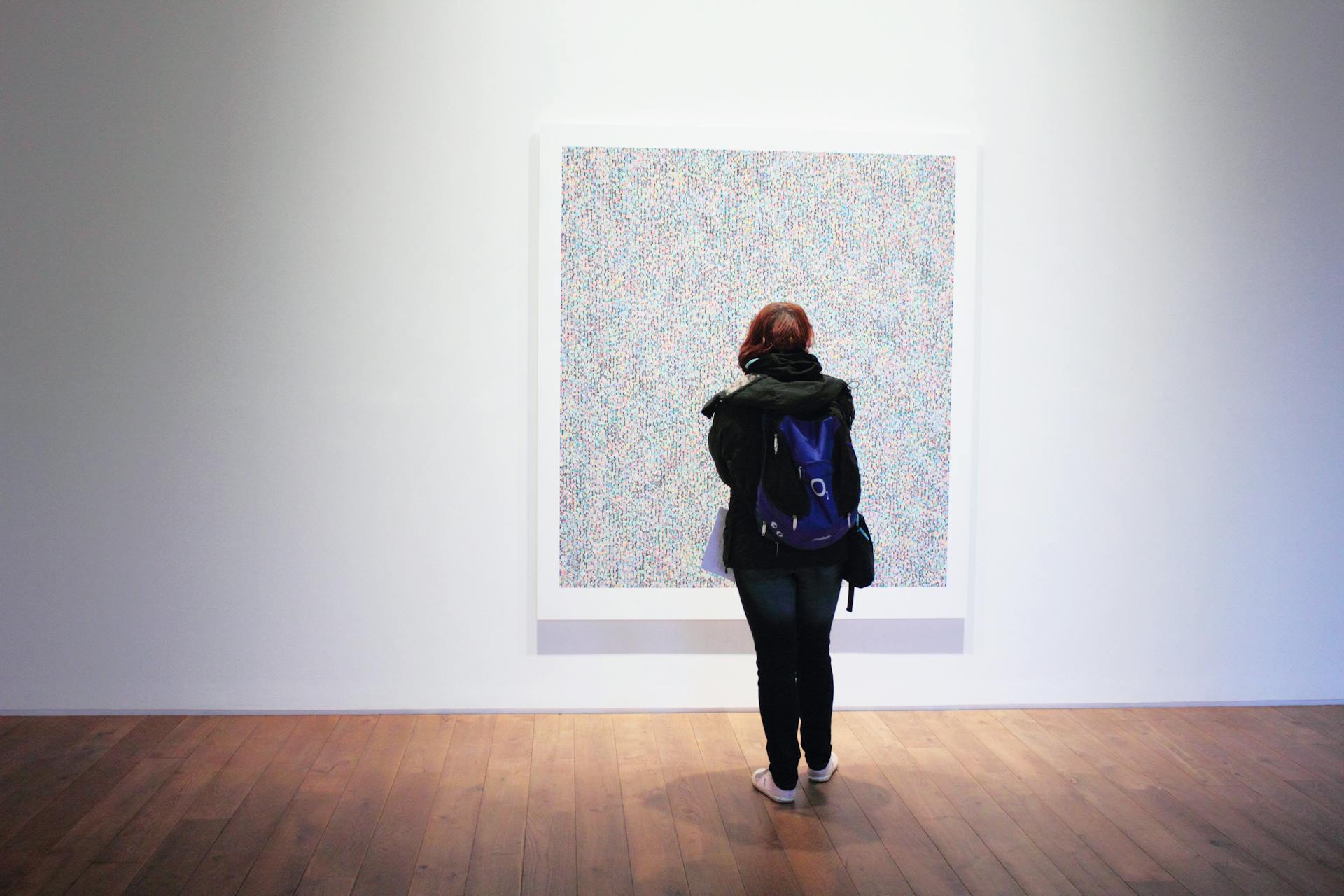 Person standing in a minimalist gallery, viewed from behind, looking at a large abstract artwork composed of dense colorful dots on a white wall.