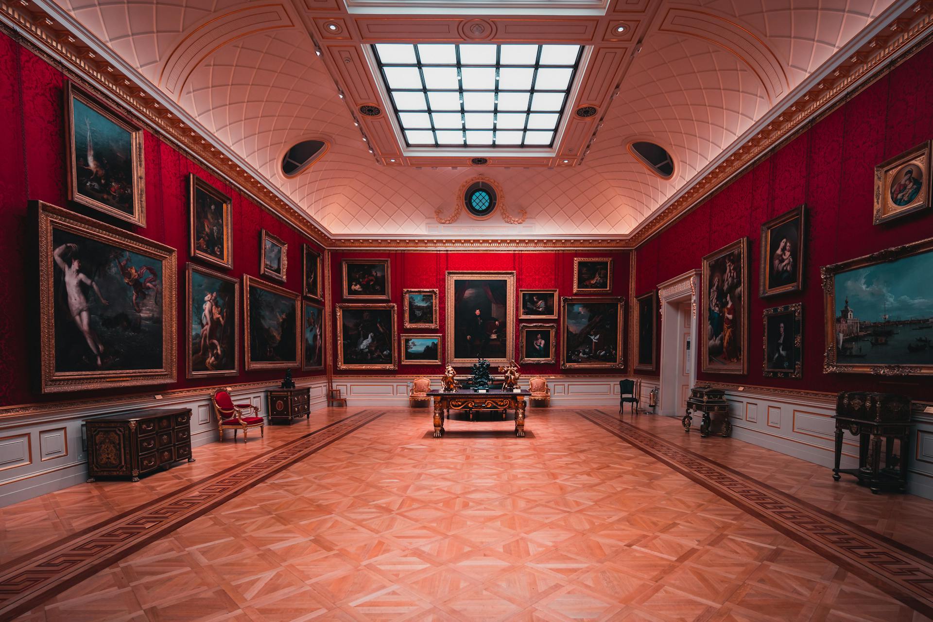 Red-walled gallery at The Frick Collection in New York, featuring classical European paintings, ornate furniture, and a skylit ceiling above a polished wood floor