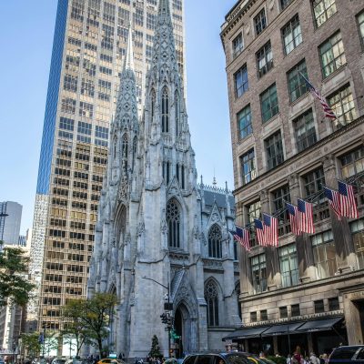 St. Patrick’s Cathedral on Fifth Avenue in Midtown Manhattan, with its Gothic spires rising between modern skyscrapers and busy New York City street traffic in the foreground