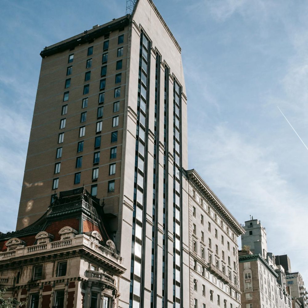 Street-level view of Upper East Side buildings in New York City, featuring a mix of modern high-rise apartments and historic architecture, including an ornate corner building with a mansard roof under a clear blue sky.