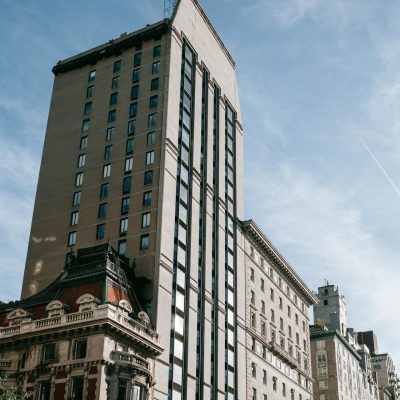 Street-level view of Upper East Side buildings in New York City, featuring a mix of modern high-rise apartments and historic architecture, including an ornate corner building with a mansard roof under a clear blue sky.
