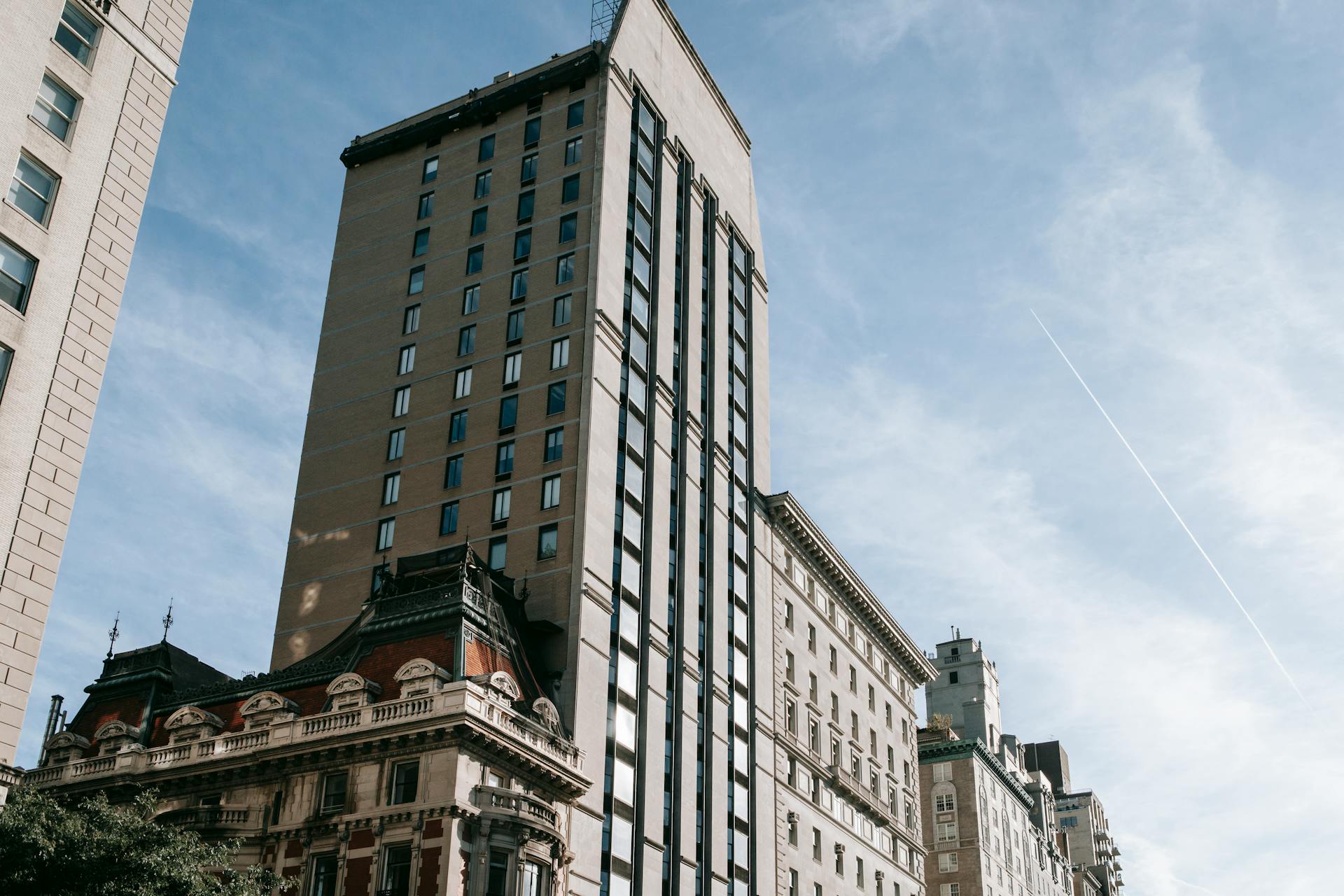 Street-level view of Upper East Side buildings in New York City, featuring a mix of modern high-rise apartments and historic architecture, including an ornate corner building with a mansard roof under a clear blue sky.
