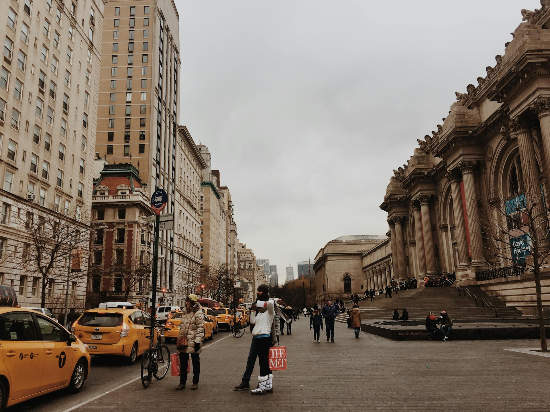 Street view along Fifth Avenue in New York City on an overcast day, with yellow taxis and pedestrians in winter clothing in the foreground, and the Metropolitan Museum of Art’s grand columned facade and steps on the right