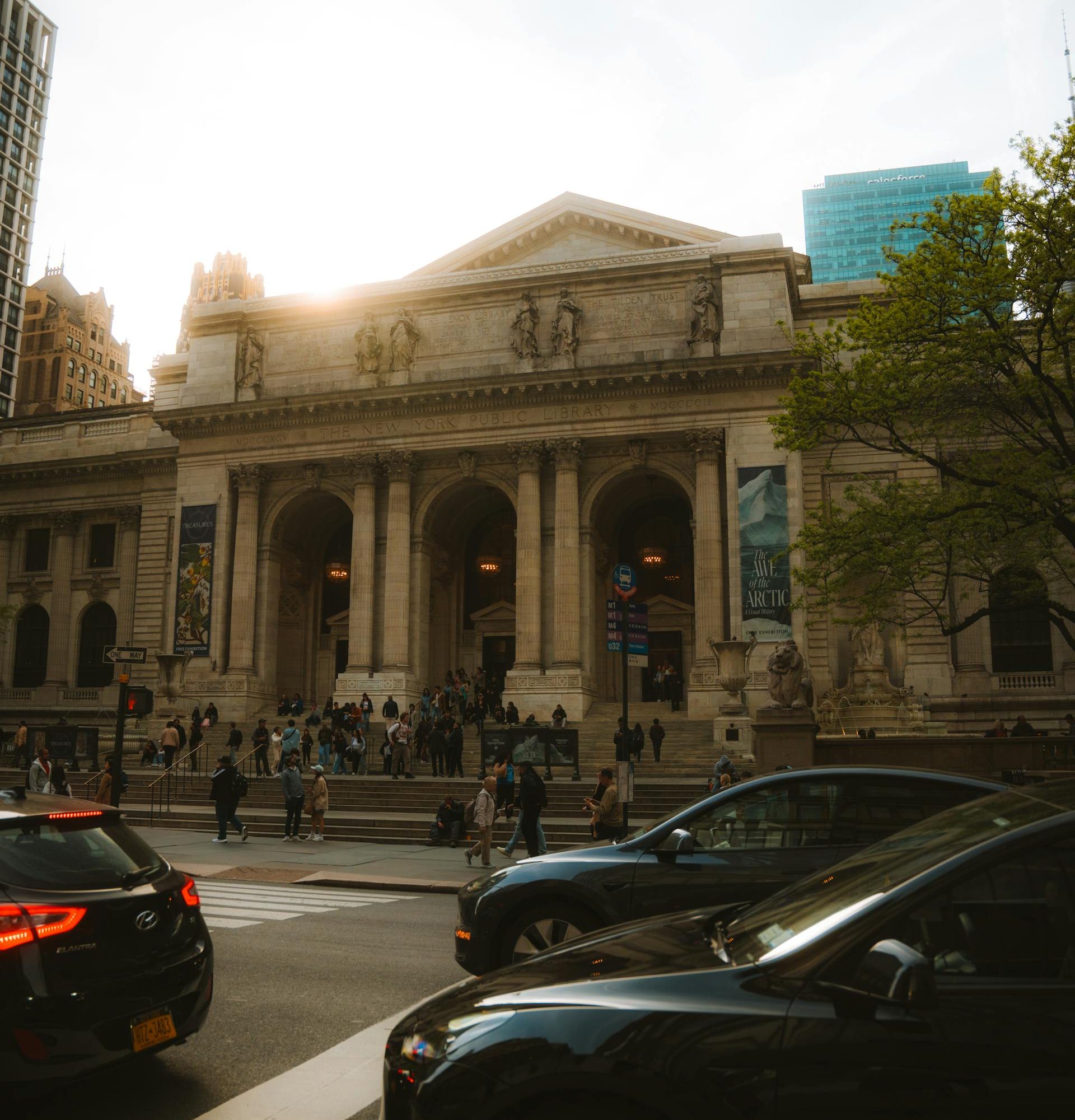 Sunlit exterior of the New York Public Library on Fifth Avenue, with people gathered on the steps and cars passing in the foreground, framed by city buildings and trees.