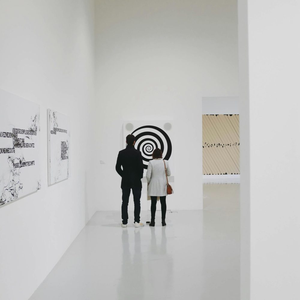 Two people standing in a bright, minimalist art gallery observing a black-and-white spiral artwork, with additional contemporary pieces displayed on surrounding white walls.